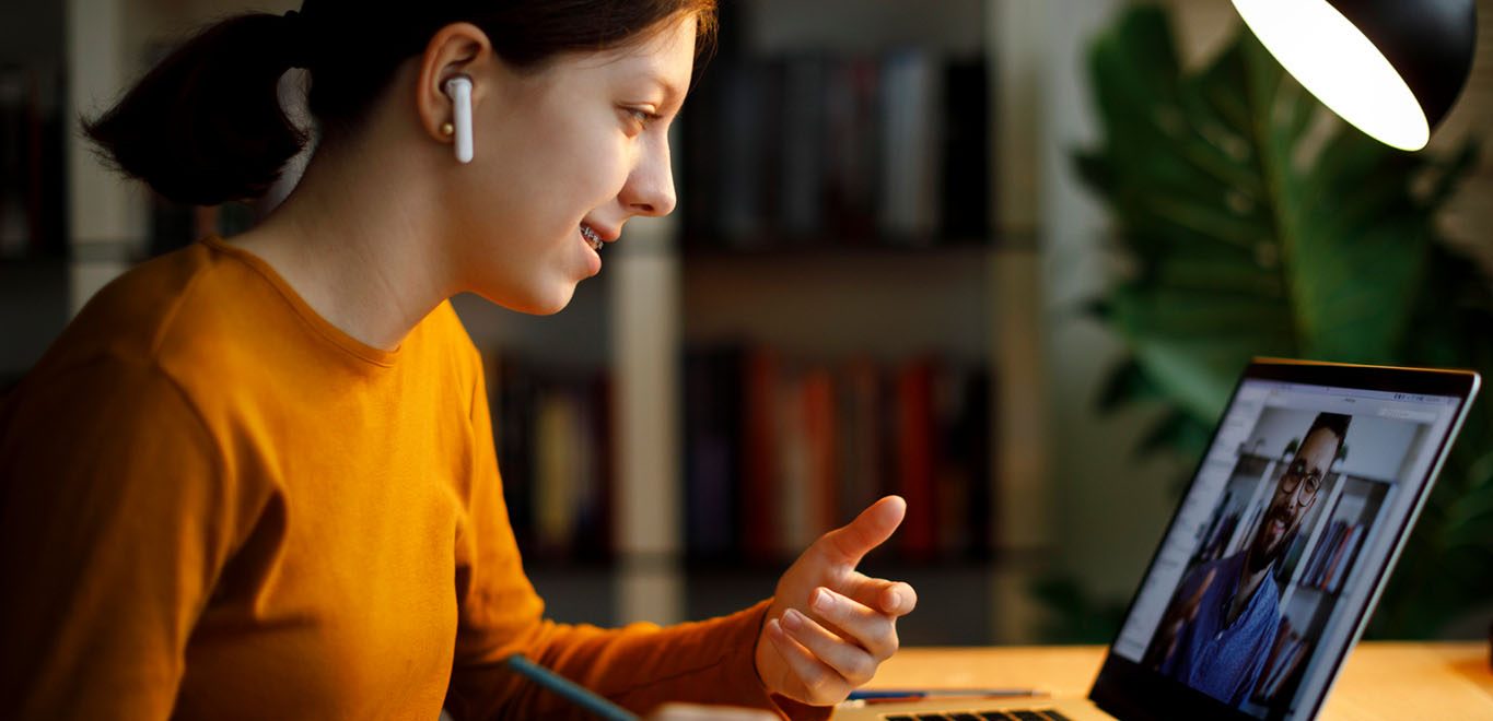 a female student wearing wireless headphones sits at a table engaged in a video conference. She is taking notes with a pencil.